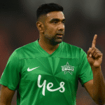 Athlete wearing green sports jersey with logo and sponsor, raising index finger during a game, symbolizing victory or alertness, on a blurred stadium background.