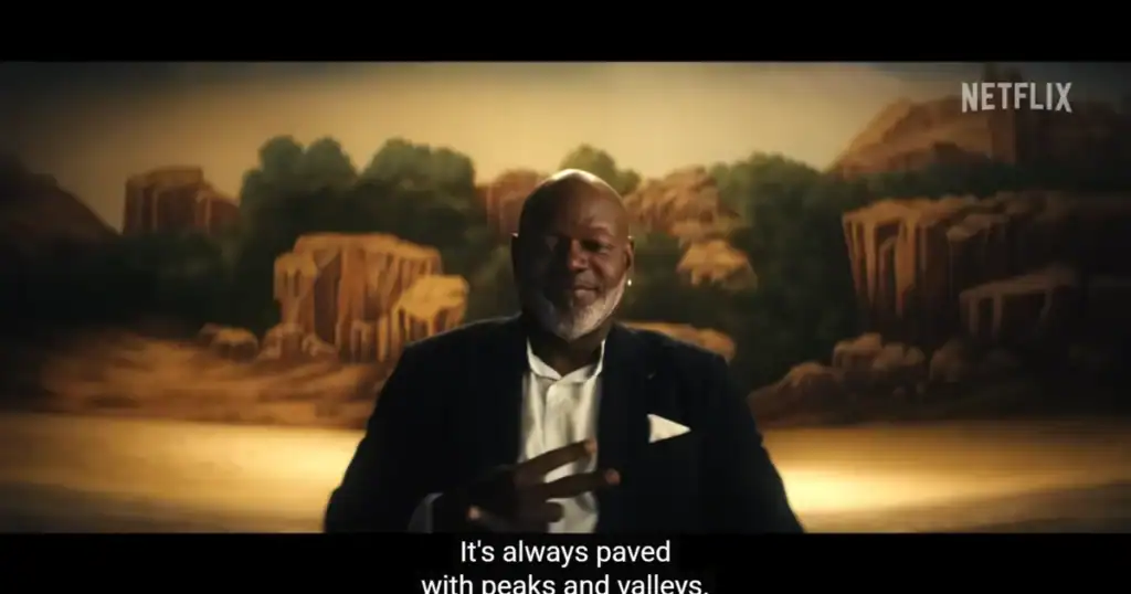 Aged man with gray beard in a suit, standing in front of a painted background of ancient ruins and lush greenery, holding a confident pose in a modern indoor setting.